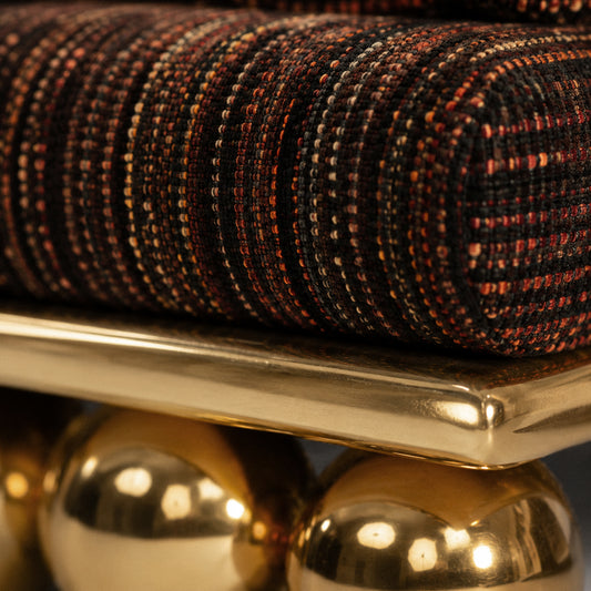 Close-up of a woven cushion or upholstered cushion cover in a dark multicolor tweed pattern, resting on a polished gold metal surface with reflective gold spheres below.