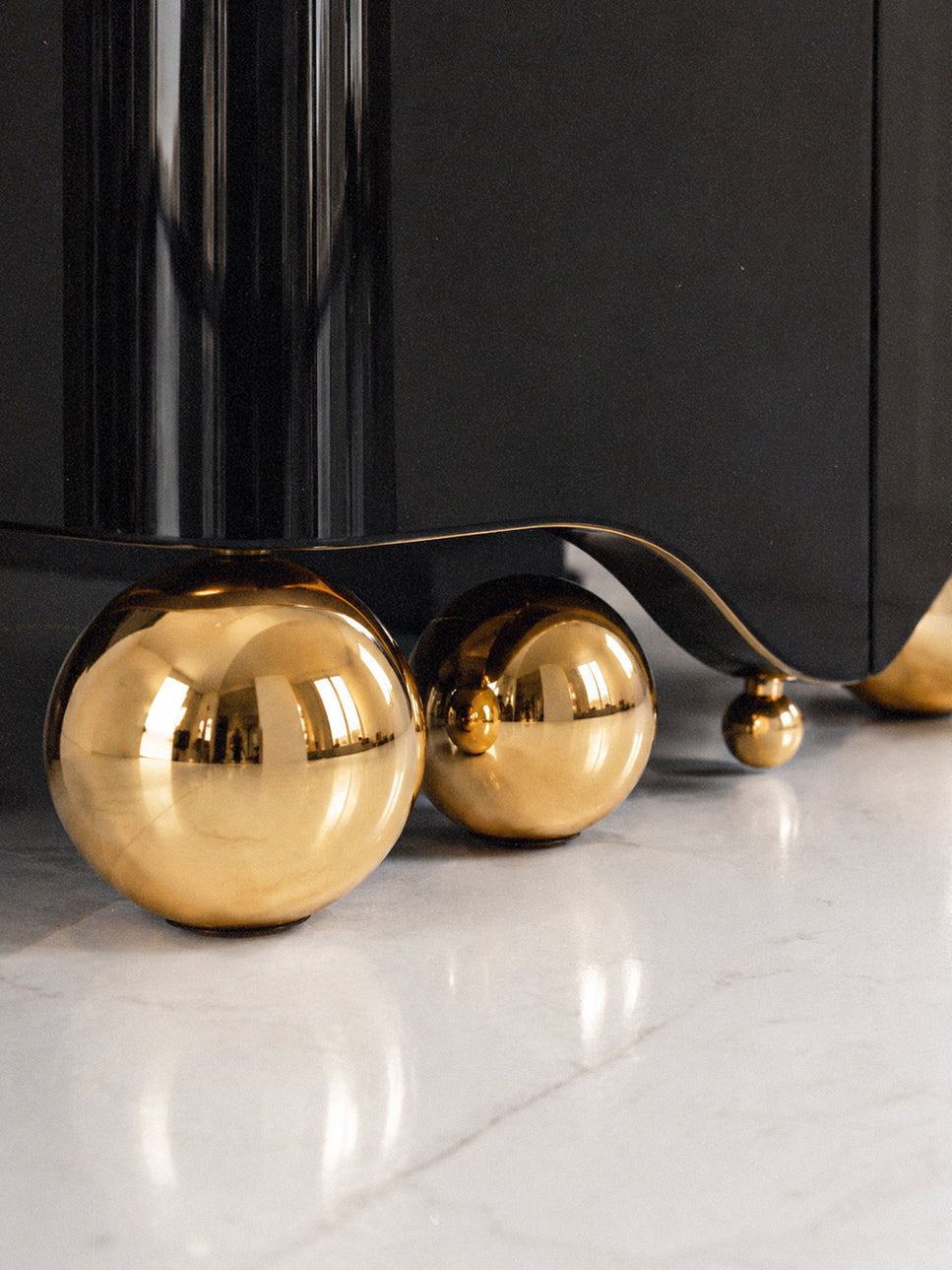 Close-up of a black cabinet or furniture piece supported by large polished gold spherical feet on a light marble floor.