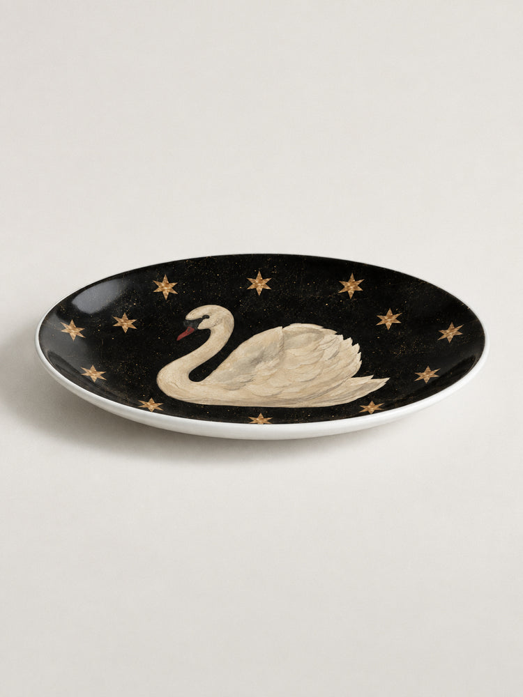 An oval black ceramic plate featuring a hand-painted white swan with a red beak, surrounded by small gold stars, shown on a neutral light beige background.
