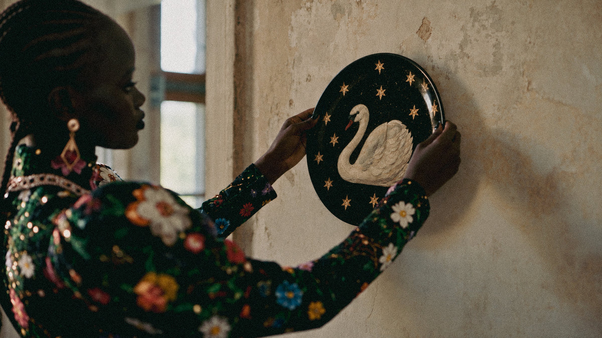 A woman in a dark floral embroidered dress hangs a round black decorative plate featuring a white swan and gold stars on a textured beige wall.