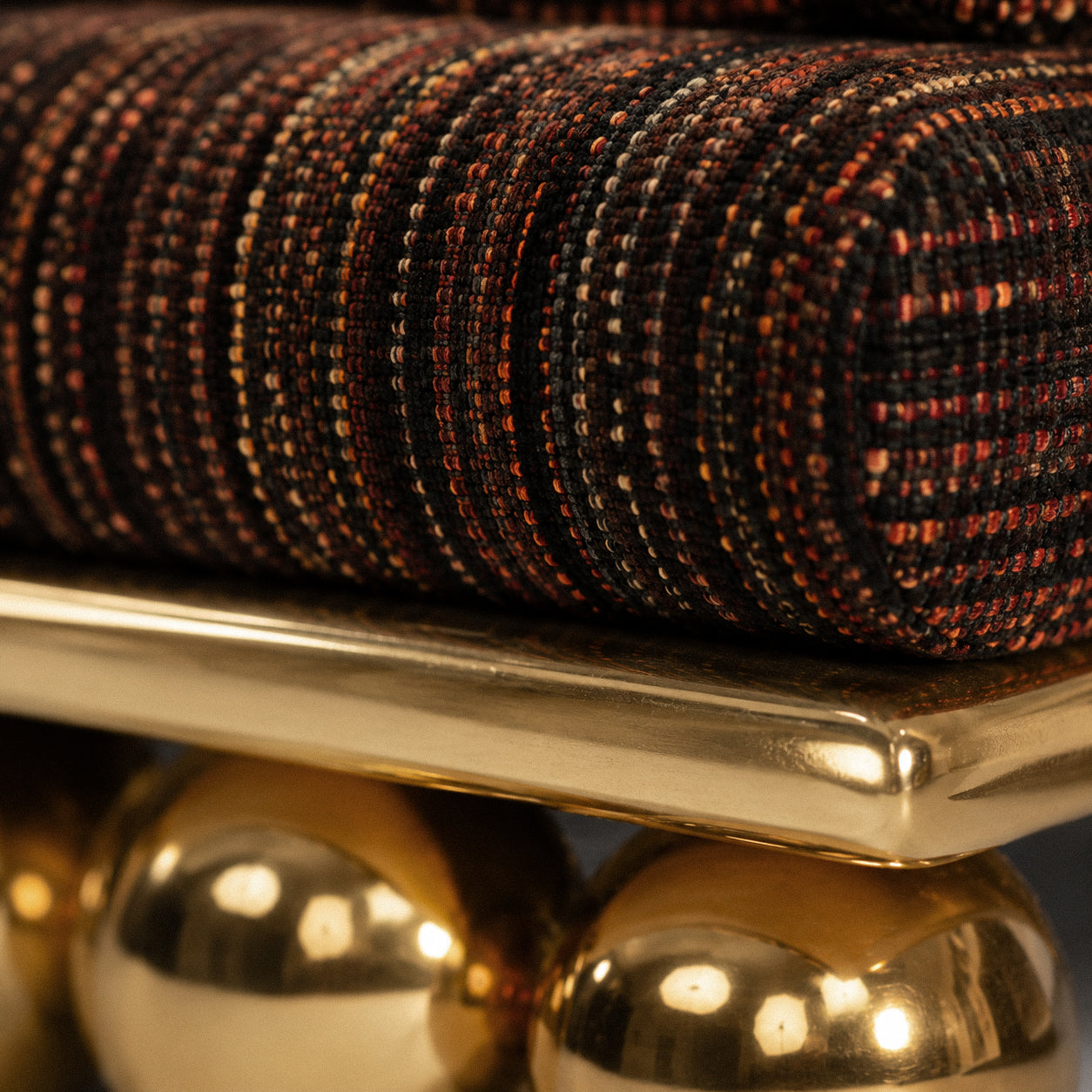 Close-up of a woven cushion or upholstered cushion cover in a dark multicolor tweed pattern, resting on a polished gold metal surface with reflective gold spheres below.