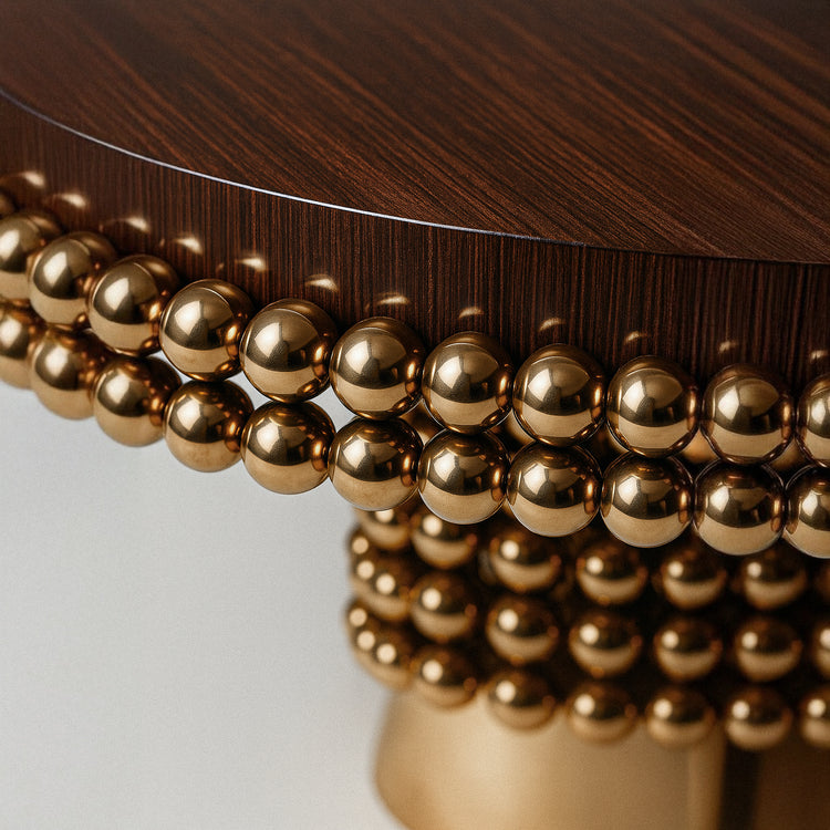 Side profile of the Lustra dining table showcasing layered polished gold spheres and a cylindrical brass pedestal base beneath a rich dark wood tabletop.