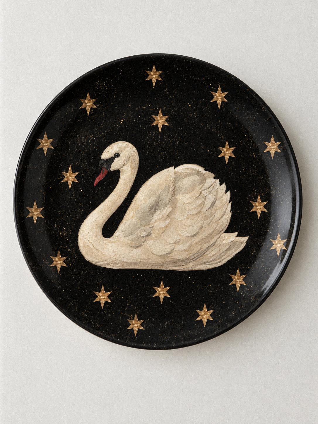 A round black decorative plate featuring a painted white swan with a red beak, surrounded by small gold stars, shown against a light neutral background.