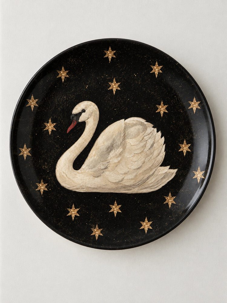A round black decorative plate featuring a painted white swan with a red beak, surrounded by small gold stars, shown against a light neutral background.